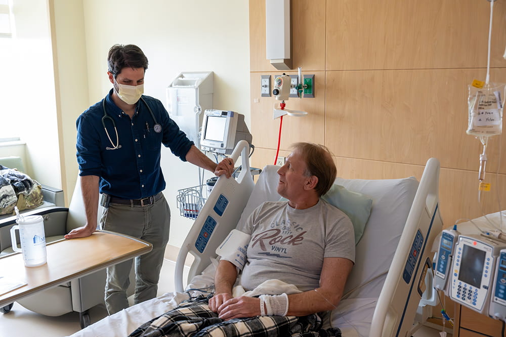 a doctor casually leans on a hospital bed in upright position to chat with the patient sitting in it 