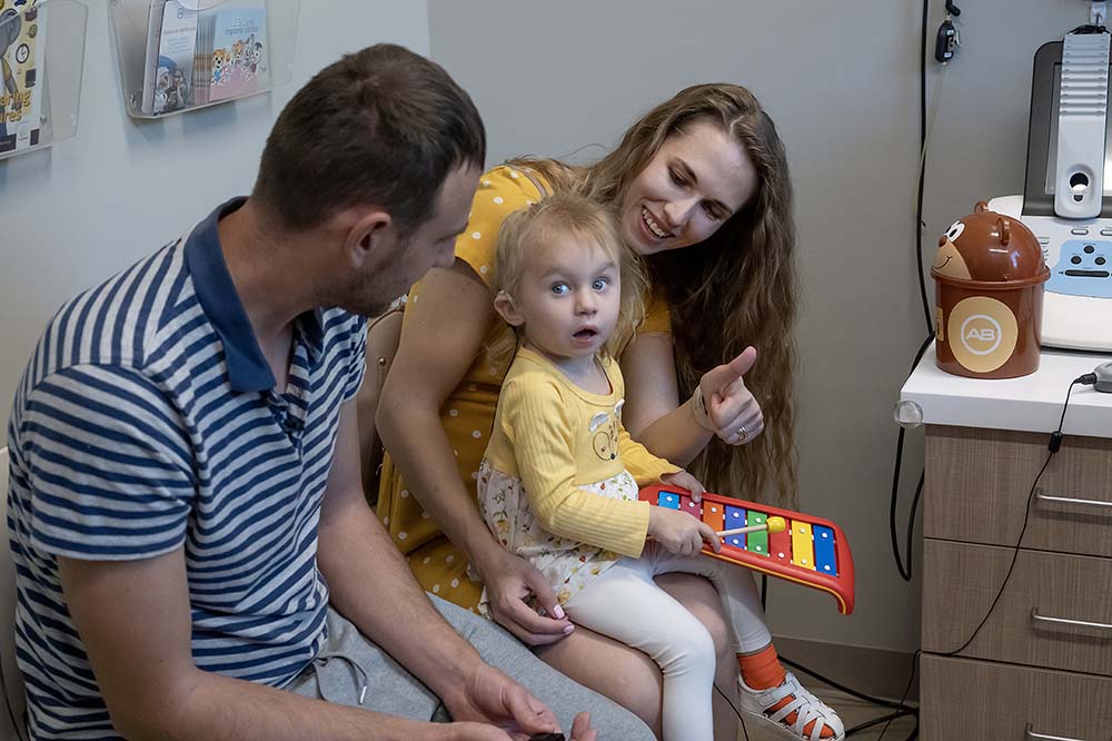 A little girl sitting on a woman's lap looks to the side. The woman is smiling and has long brown hair. A man wearing a striped shirt sits beside them. He also has brown hair.