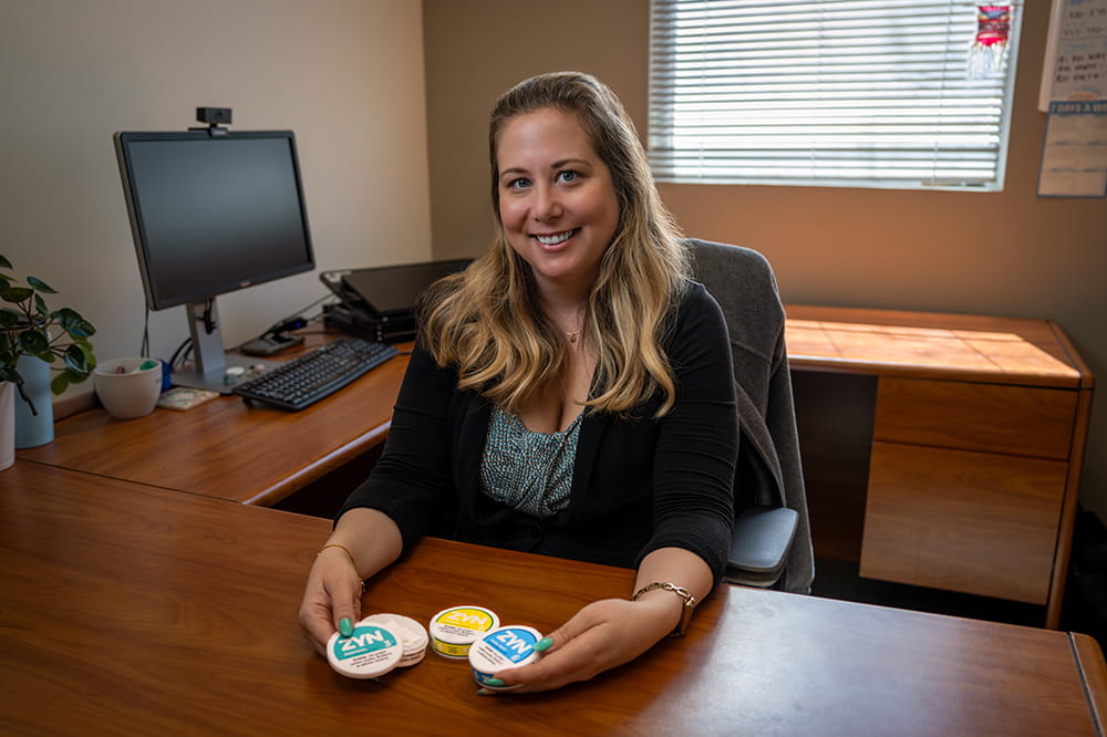 a woman sits at a desk and shows Zyn tins